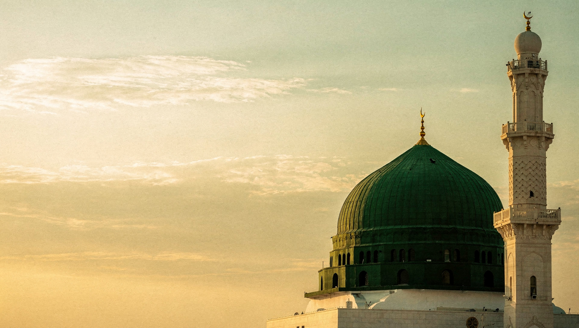 The Green Dome in Madinah at dawn, beside a minaret, under a soft gold sky.